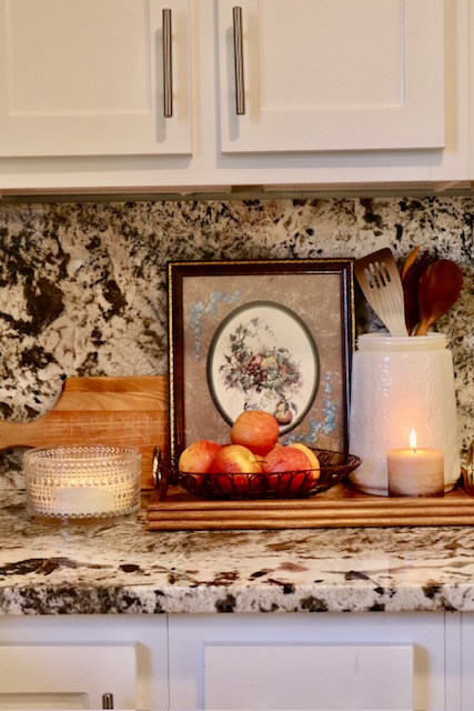 Kitchen counter display with a framed floral picture, a wire bowl of apples, wooden utensils in a white jar, a lit candle, and a small cutting board against a marble backsplash.