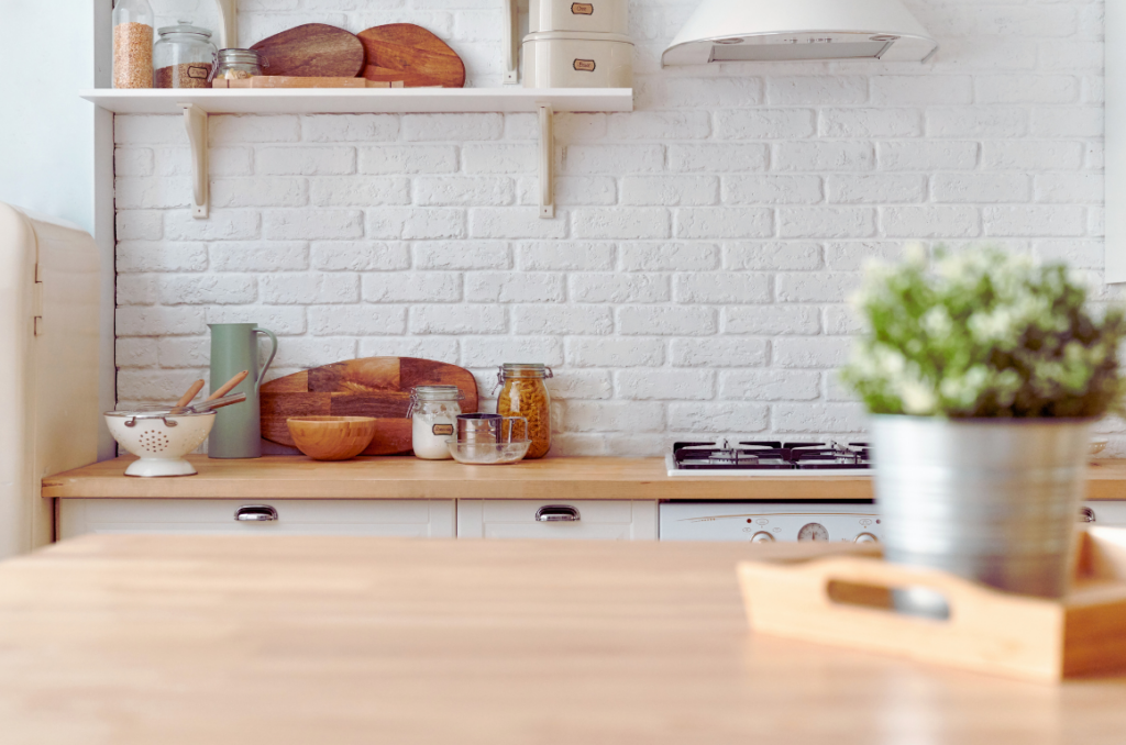 A bright kitchen with a wooden countertop, white brick wall, open shelving, jars, cutting boards, and a metal plant pot on a wooden tray in the foreground.